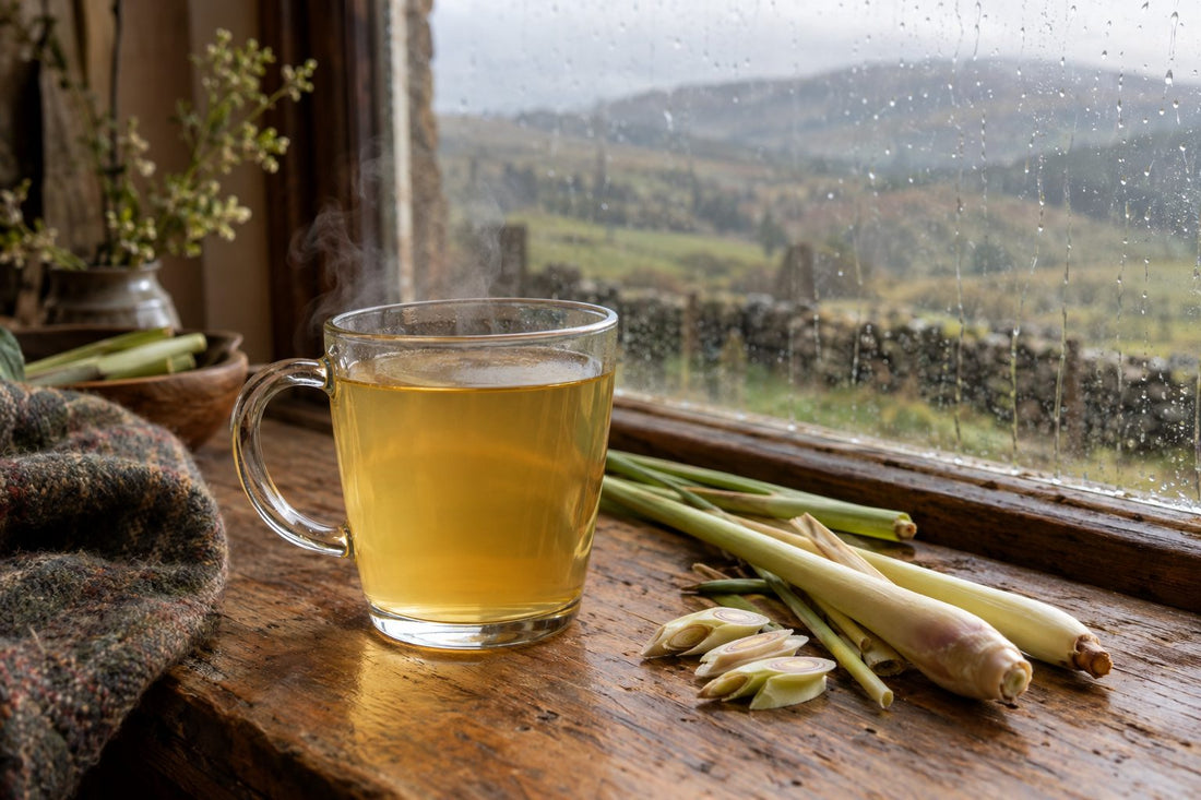 Glass mug of pale golden lemongrass tea with fresh stalks on an oak table by a rainy window
