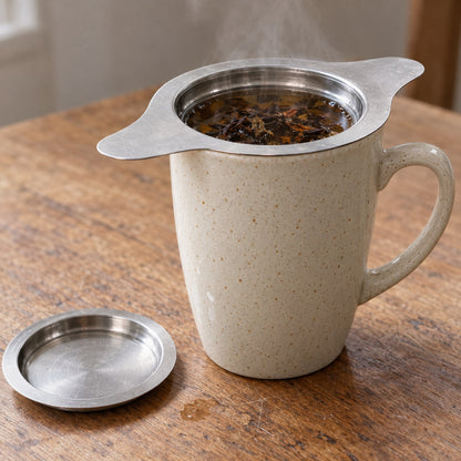 Basket infuser sitting in a stoneware mug with its wings resting on the rim, black tea brewing, lid on the table