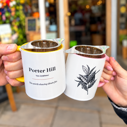 Yellow and pink Porter Hill mugs with basket infusers held outside the tea shop, showing the logo and botanical tea leaf illustration