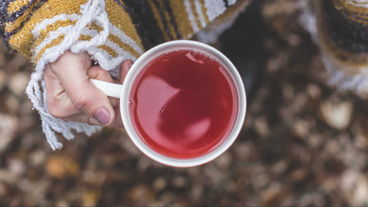Person holding a mug of red liquid wrapped in a patterned blanket outdoors.
